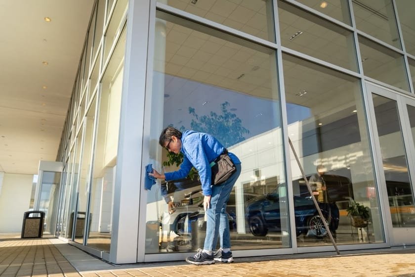 A window cleaner in a blue jacket wiping the edge of a large showroom window by hand