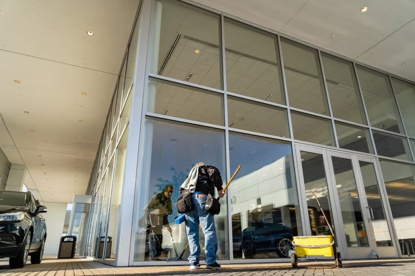 A window cleaner with a bucket cart and squeegee pole working along the floor-to-ceiling glass of a car showroom