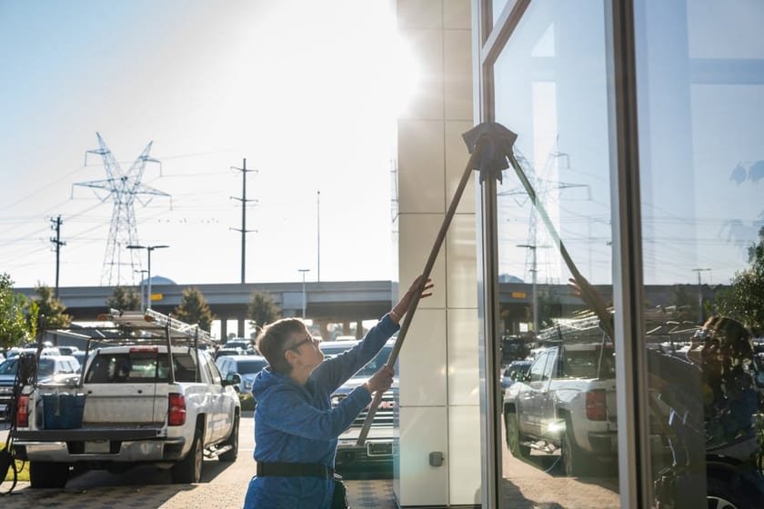 A window cleaner reaching up with an extension pole to the top of a tall glass panel, pickup trucks parked behind