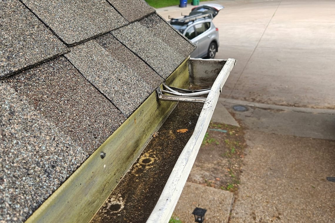 An open gutter holding silt and debris along the edge of a shingled roof